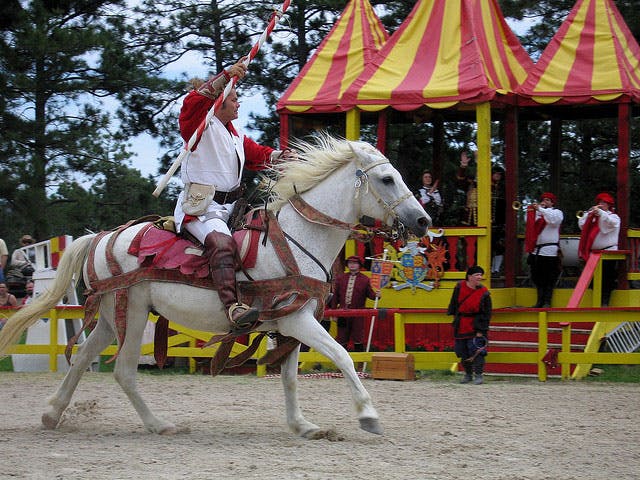 A colorful jouster and his horse at the Colorado Renaissance Festival with red and yellow tents behind and lots of spectators.