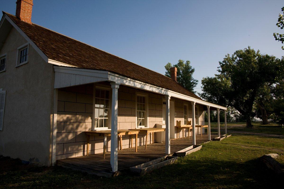 A wooden house sits on the left with two chimneys and tables on the porch. On the right green grass has green-leafed trees.