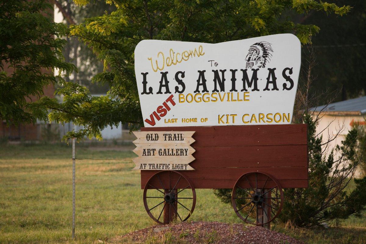 A sign that looks like a covered wagon reads "Welcome to Las Animas Visit Boggsville Last Home of Kit Carson" on the cover part. On the wooden carriage is a wooden sign that reads "Old Trail Art Gallery at Traffic Light." Behind the sign is an evergreen tree and green grass.