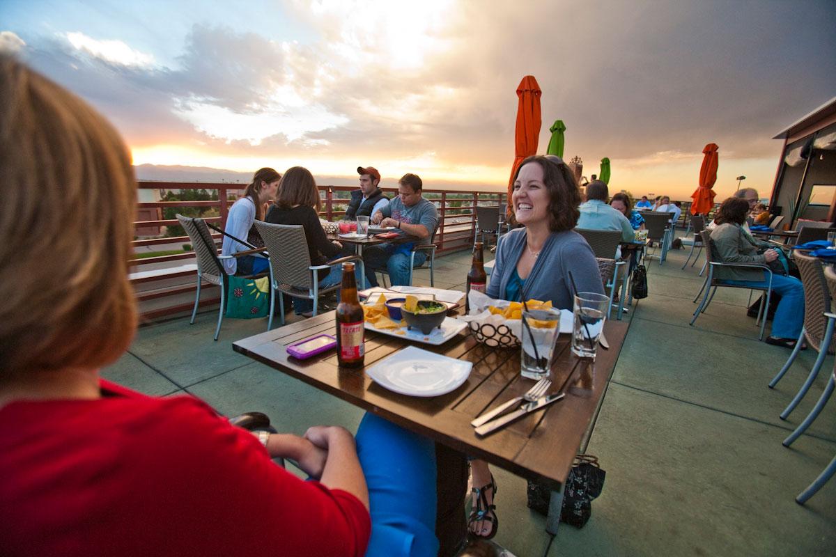 A woman smiles across the table at her dining companion, they on an outdoor rooftop, where the sun sets and other diners sit at tables behind them