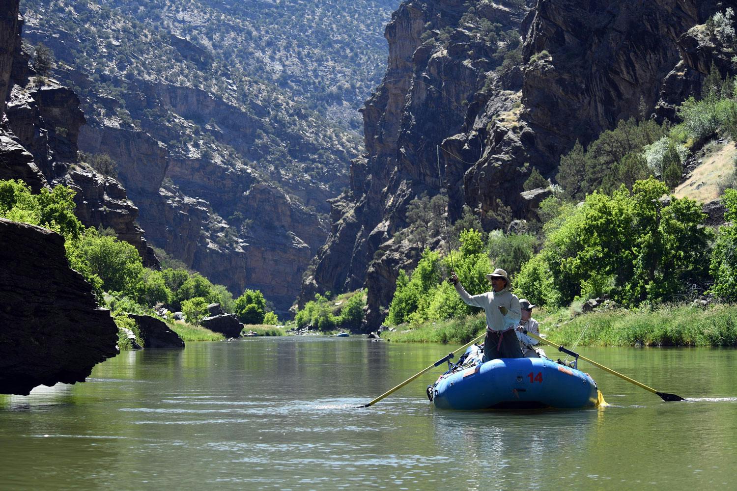 An inflatable blue raft with two people in it sits in the middle of the calm Gunnison River surrounded by vegetation and rocky walls.