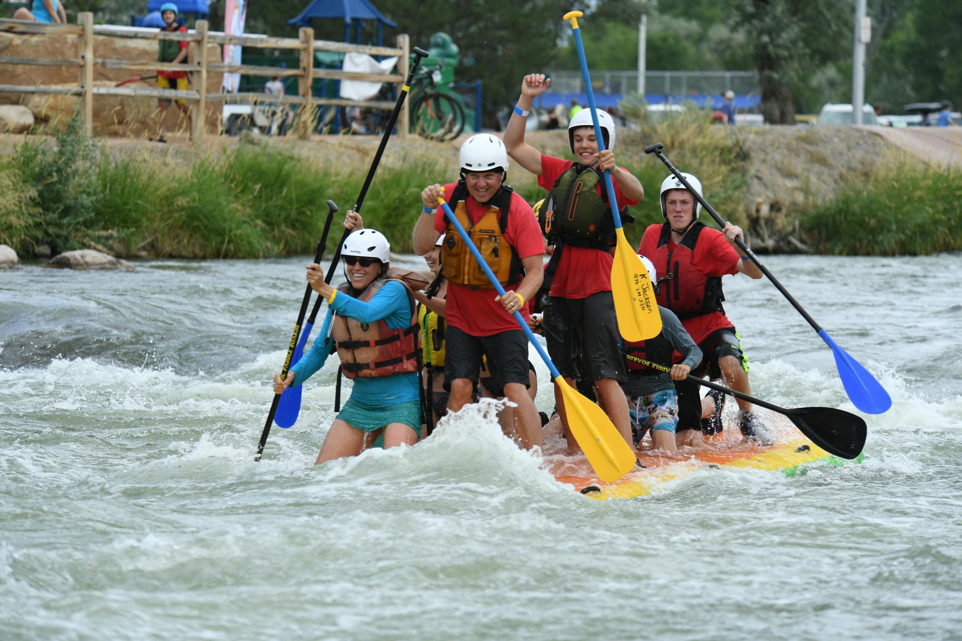 More than four people in white helmets and life jackets stand atop a floating raft in the middle of a rushing water at the Montrose Water Sports Park.