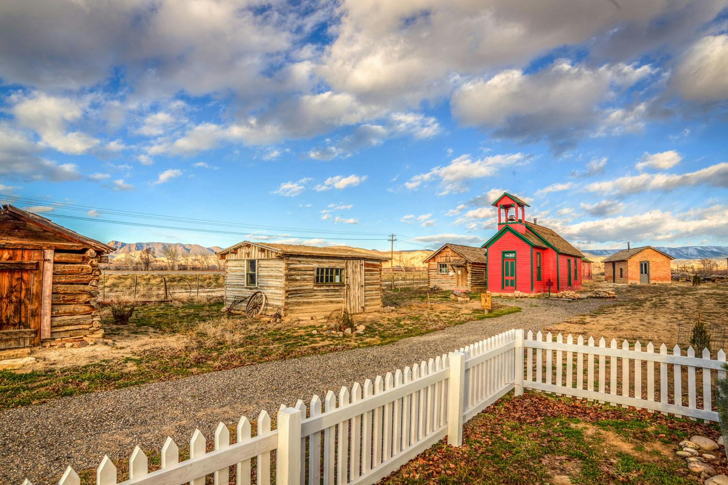 Small log cabins with a bright-red building sit in yellow and green grass with mountains in the background under a blue sky with clouds.