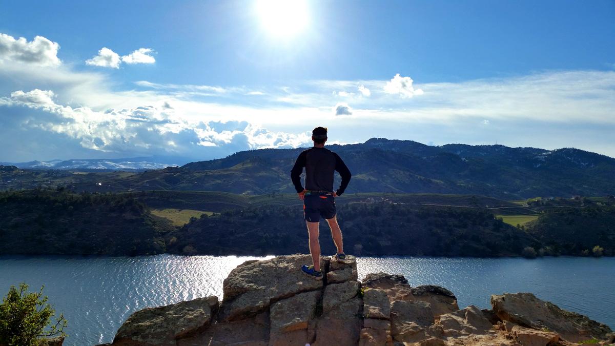 A person stands on a rock viewpoint looking out at the rippling, blue Horsetooth Reservoir. In the distance green grass, and trees roll into mountains. The sky is blue, the sun is a bright ball and there are white clouds.