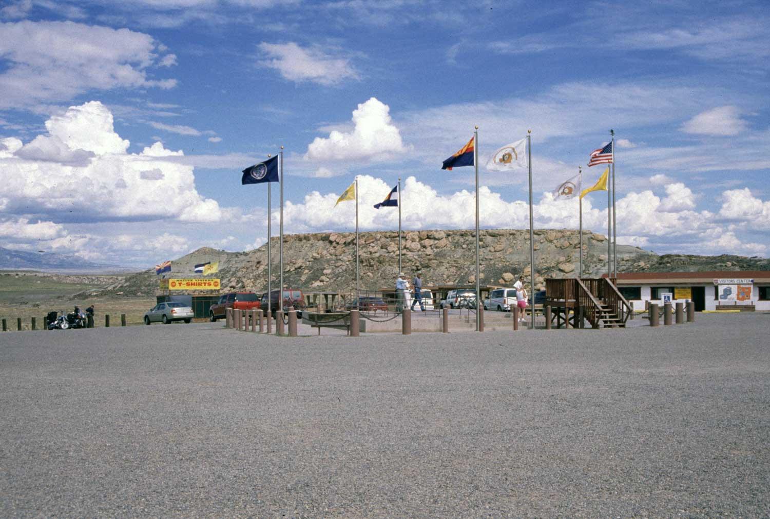 Several flags fly in the wind atop tall flag poles at the Four Corners Monument in southwest Colorado. The sky above is a vibrant blue and is spotted with fluffy clouds.