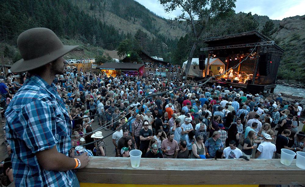 A person in a brimmed hat leans against a railing overlooking the crowd watching a musical group on the stage of the open-air Mishawaka Amphitheater in Fort Collins, Colorado