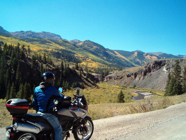 A motorcyclist in a blue jacket and jeans with a black helmet on sits on a dirt pullout looking at the view. The view has a blue sky, mountains covered in evergreen trees and golden aspens.