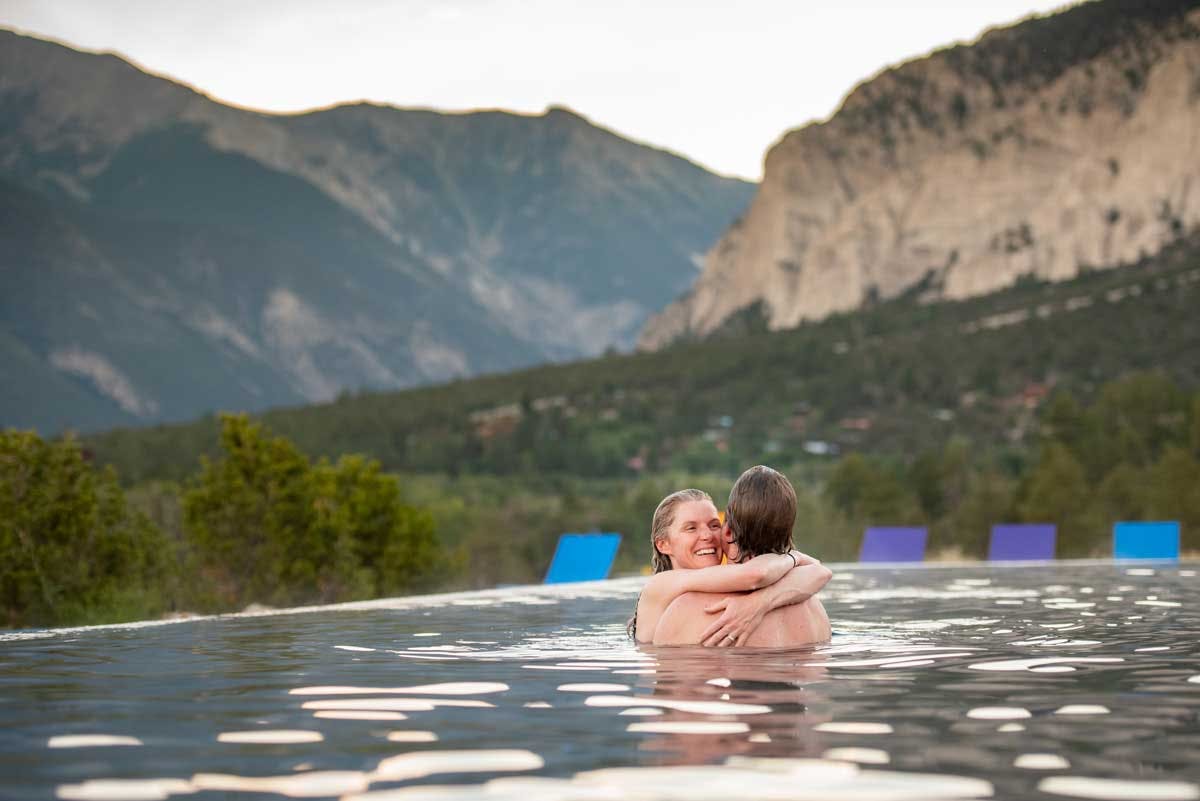 A couple embraces in a pool at Mount Princeton Hot Springs Resort. There are rock-faced buttes with evergreen trees in the background.