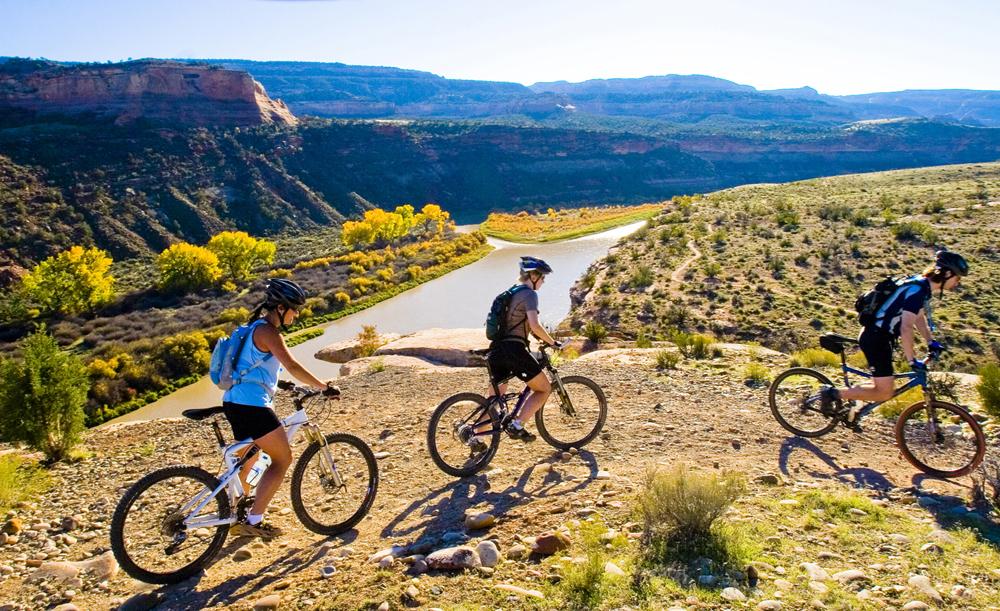 A group of bikers go around a bend in the path near Fruita. The bend overlooks a flowing river with foliage.