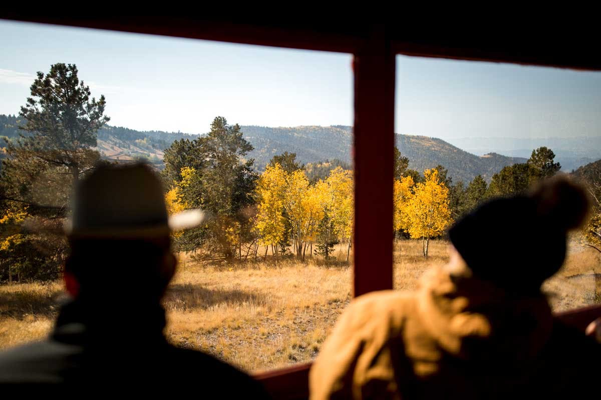 Two people sit in a car of the Cripple Creek and Victor Narrow Gauge Railroad looking out an open window at a late-afternoon fall scene. The grass is yellow, the trees are turning golden and there are evergreens on the left with a mountain in the background.