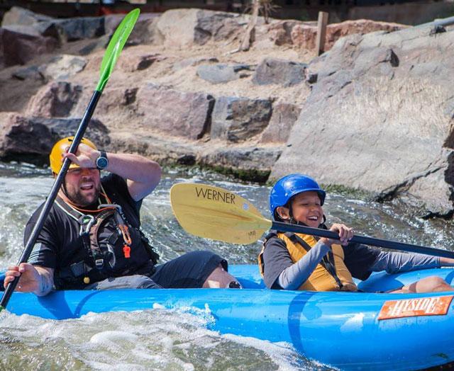 An adult and a child wear helmets and paddle through whitewater rapids in a blue raft on a Colorado river.