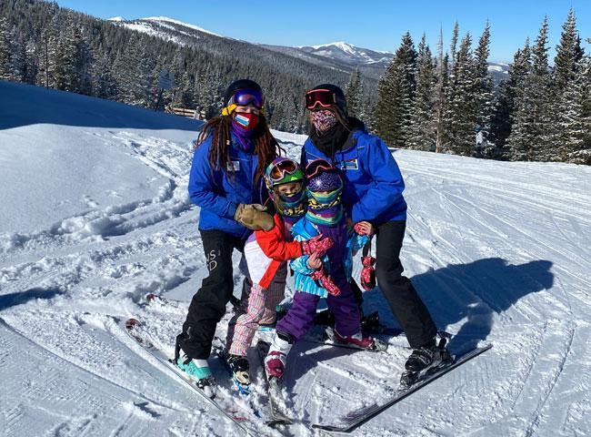 Two ski instructors in blue jackets and two children learners pose for a picture on the slopes in Colorado.