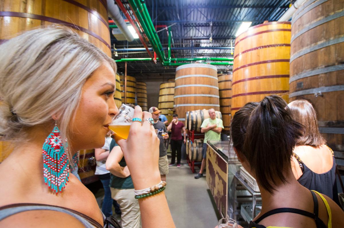 A group of people surrounded by large wooden beer barrels that reach the ceiling sip beer from small tasting glasses at a brewery in Fort Collins, Colorado.