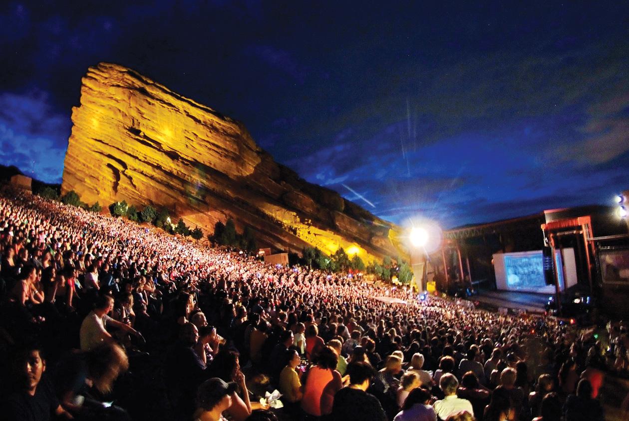 A nighttime concert takes place at Red Rocks. The stands are full, facing the stage with the red-rock geologic formation in the background.