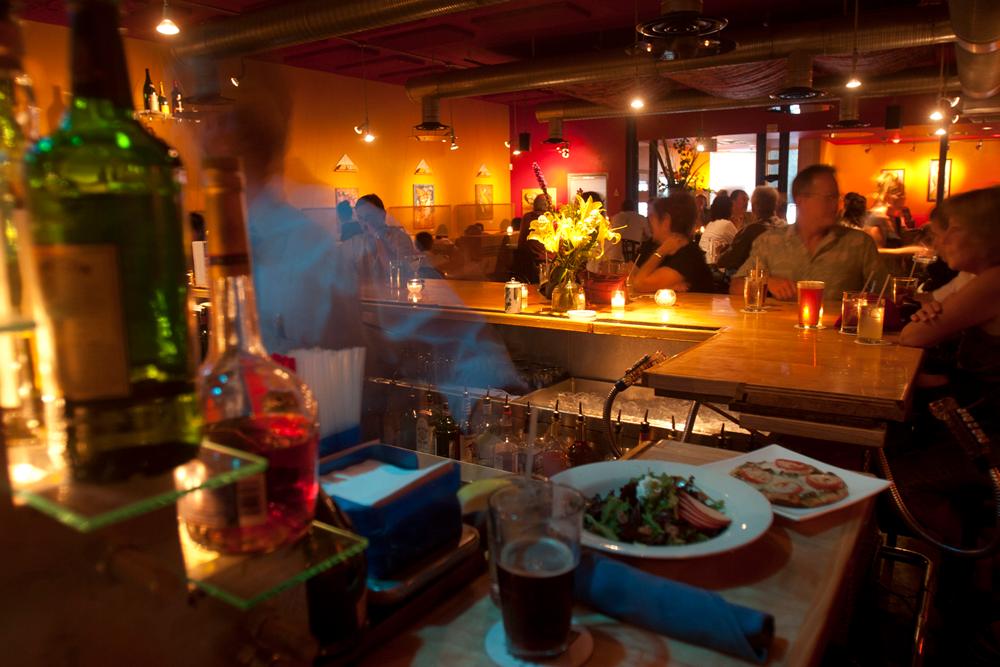 The view from behind the bar of a restaurant shows two dishes and a beer waiting to be taken to their tables; bar patrons sit in front of their beers and cocktails