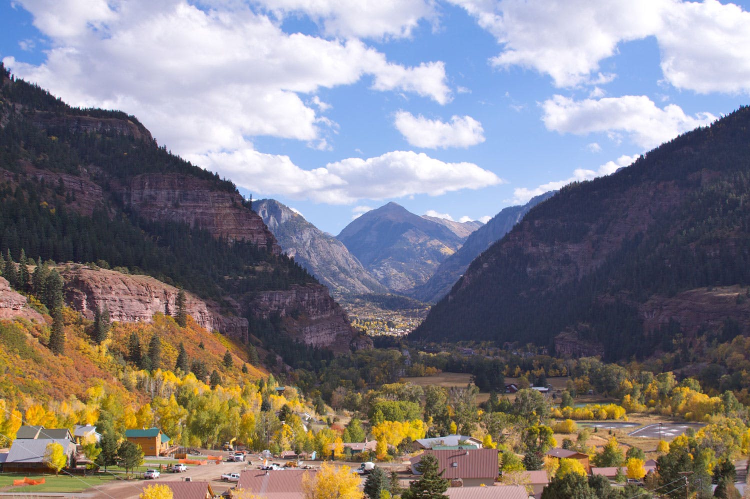 An aerial view of the town of Ouray with golden aspens and green trees. In the distance rock-mountain walls rise up in the distance. The sky is blue with clouds.