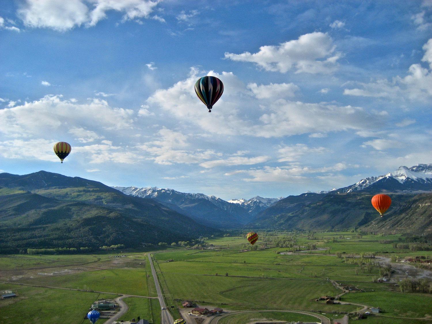 Four hot air balloons rise up into a blue, cloudy sky over a green-grass field with mountains in the distance.