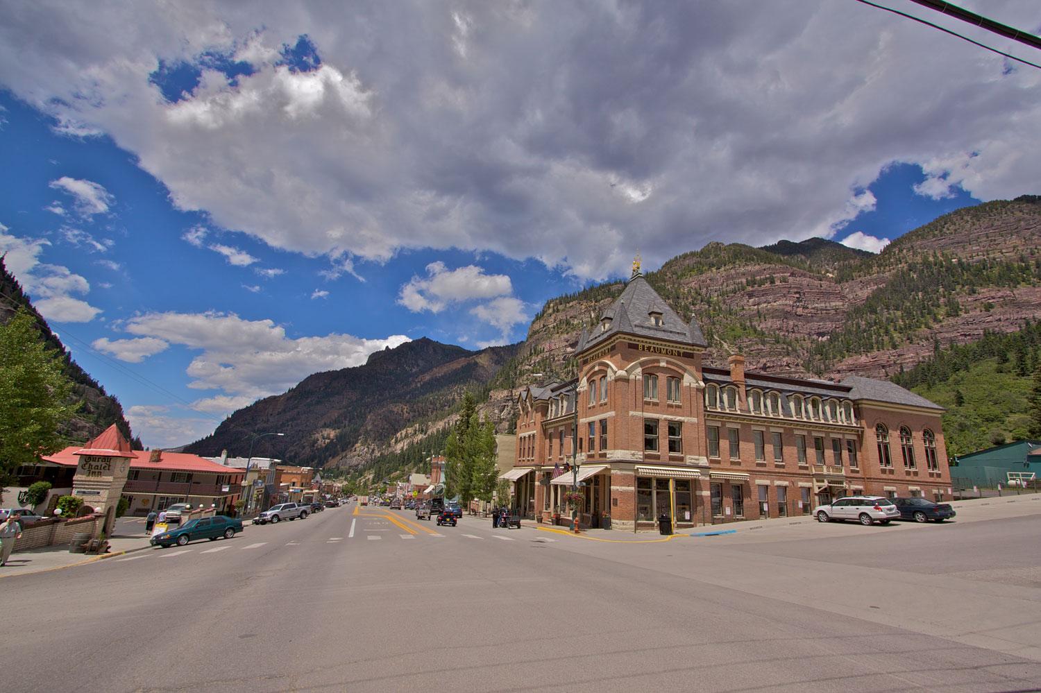 Downtown Ouray with a brick Richardson-Romanesque building on one corner and a red-roofed building on the other. In the background mountains covered in green trees with exposed stone walls rise up in a blue sky with white clouds.