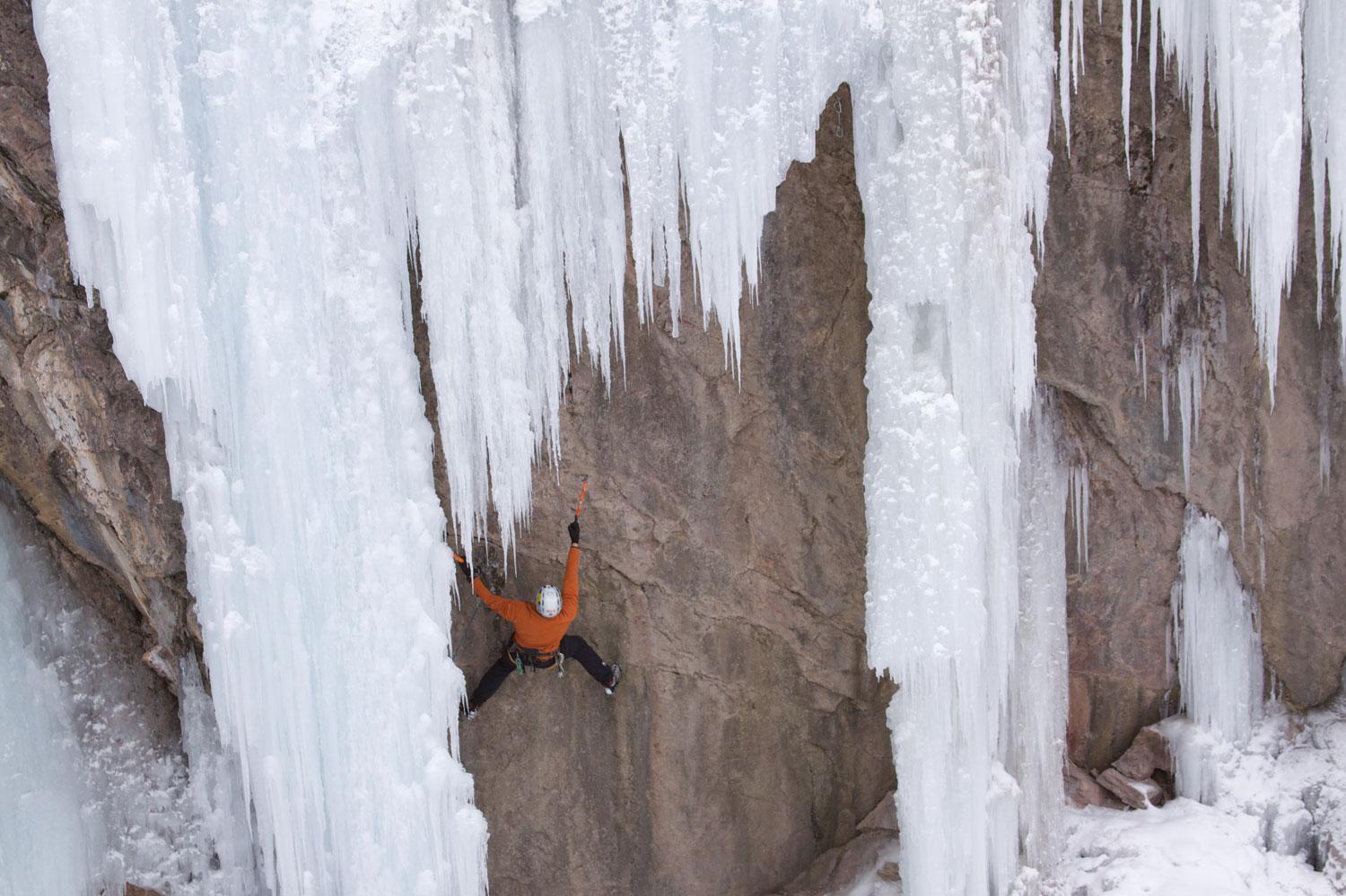 A solo ice climber climbs up a rock face with frozen, white ice structures. 