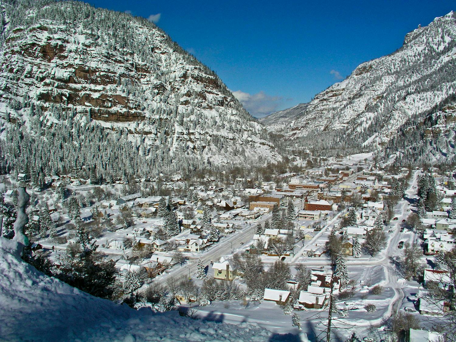 A snow-covered Ouray from above with snow-dusted mountains with exposed rock faces and evergreen trees sit under a blue sky.