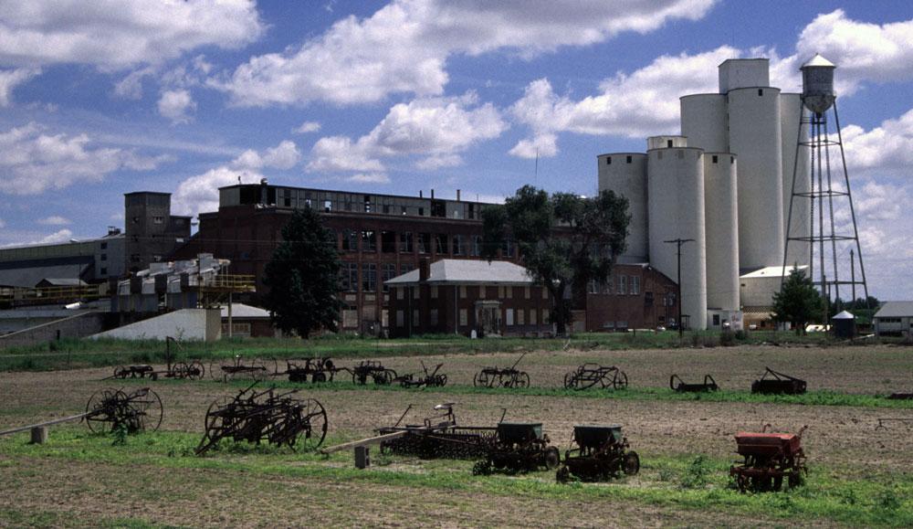 Old-fashioned farm equipment rests in two lines of grass in front of a massive sugar beet mill in Ovid, Colorado.