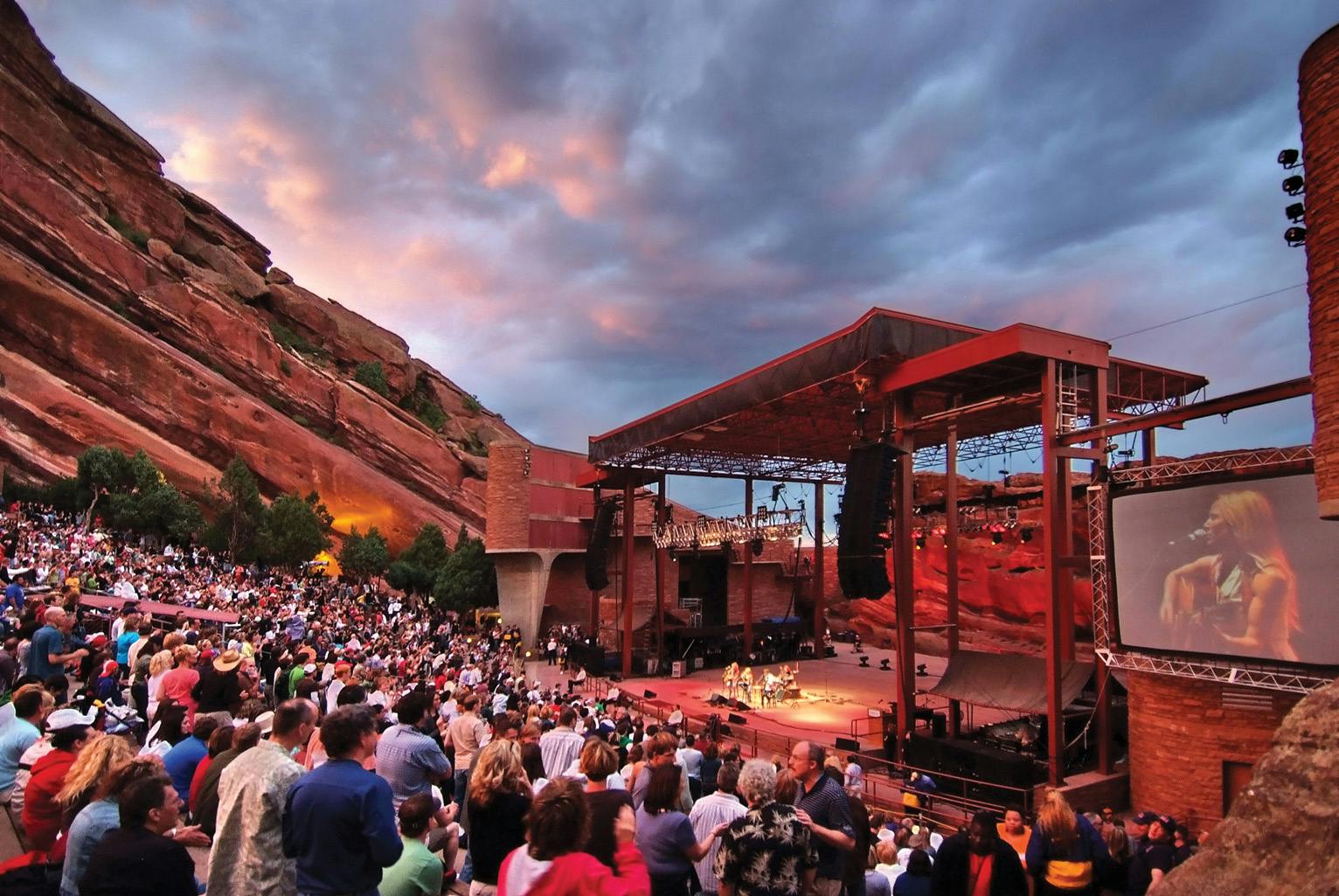 A packed concert at Red Rocks at dusk, the sky is pink and grey and the red-rock geologic formations protrude in the background.