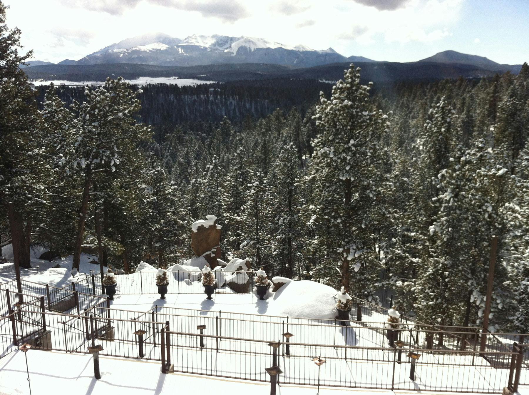 The formidable Pikes Peak is in the background of this snow-covered scene. The evergreen trees are dusted with snow and in the foreground there's an iron fence of a bed and breakfast.