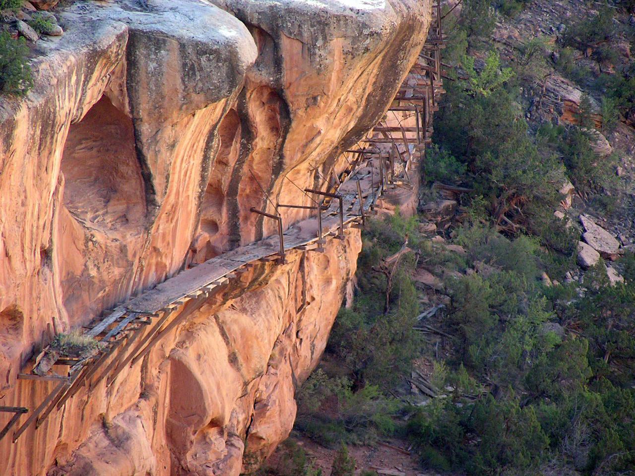 The relics of a hanging flume hug the edge of a red-rock canyon. On the left scrubby green bushes with boulders.