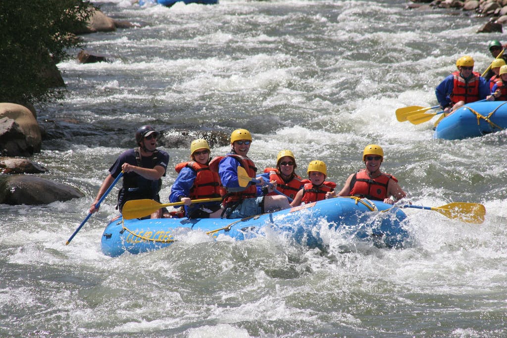 An instructor and and five paddlers in yellow helmets ride whitewater rapids in a blue raft on Clear Creek near Idaho Springs, Colorado.