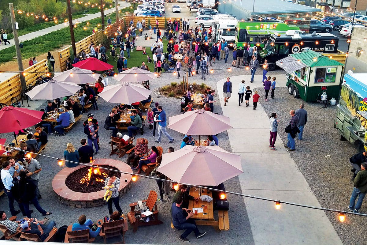 Strings of lights hang over a gravel courtyard filled with food trucks, umbrella-covered tables and a large firepit in Boulder, Colorado. People mingle, chat and eat food.