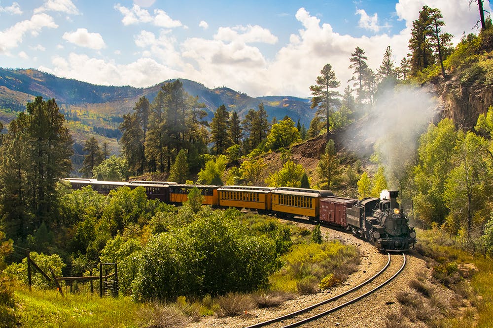 The Durango & Silverton Narrow Gauge Railroad makes its way through green vegetation on a bend in the track. In the distance there are mountain peaks under a light blue sky with white clouds.