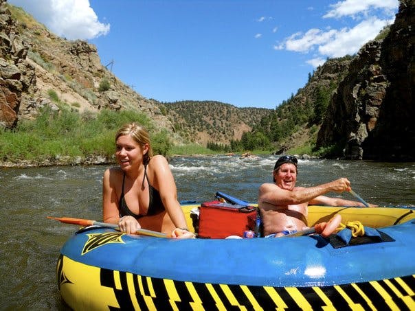 A couple sits in a blue-topped yellow and black striped tube in the middle of the Colorado River on a summer's day. The rocky walls of the canyon are covered in green tufts of grass.