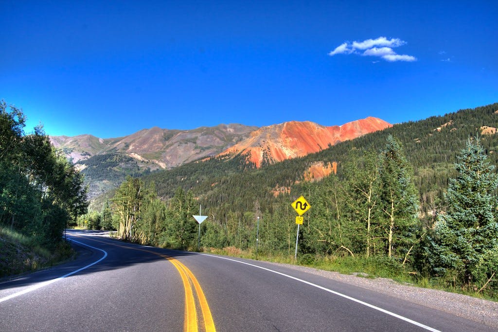 A scenic road with green trees on both sides and a red-rock mountain in the background