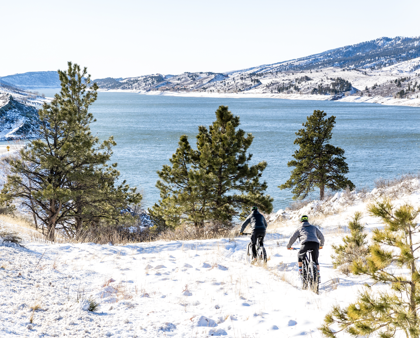 Two people ride fat bikes on a snowy trail overlooking a blue reservoir with evergreen trees in Fort Collins, Colorado.