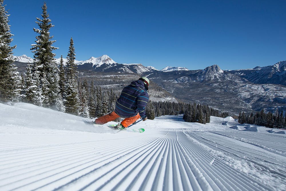 A person in orange pants and a blue jacket skis down fresh corduroy on a blue-bird day with evergreen trees dusted with snow on the left. There are mountain peaks covered in snow in the distance.