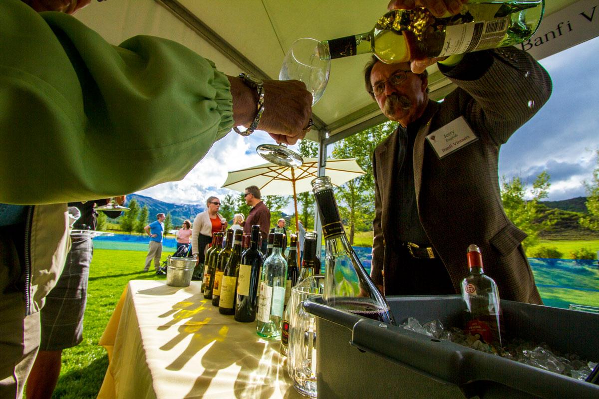 On a sunny day in Snowmass, Colorado, lights shines into a tent onto several bottles of wine. In the tent, a person pours wine into an outstretched glass.