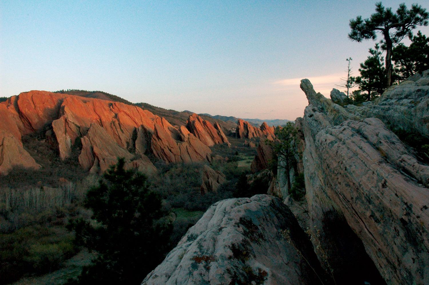 The sun sets, casting the jagged red outcroppings of the park to slowly fall into shadow, surrounded by green trees