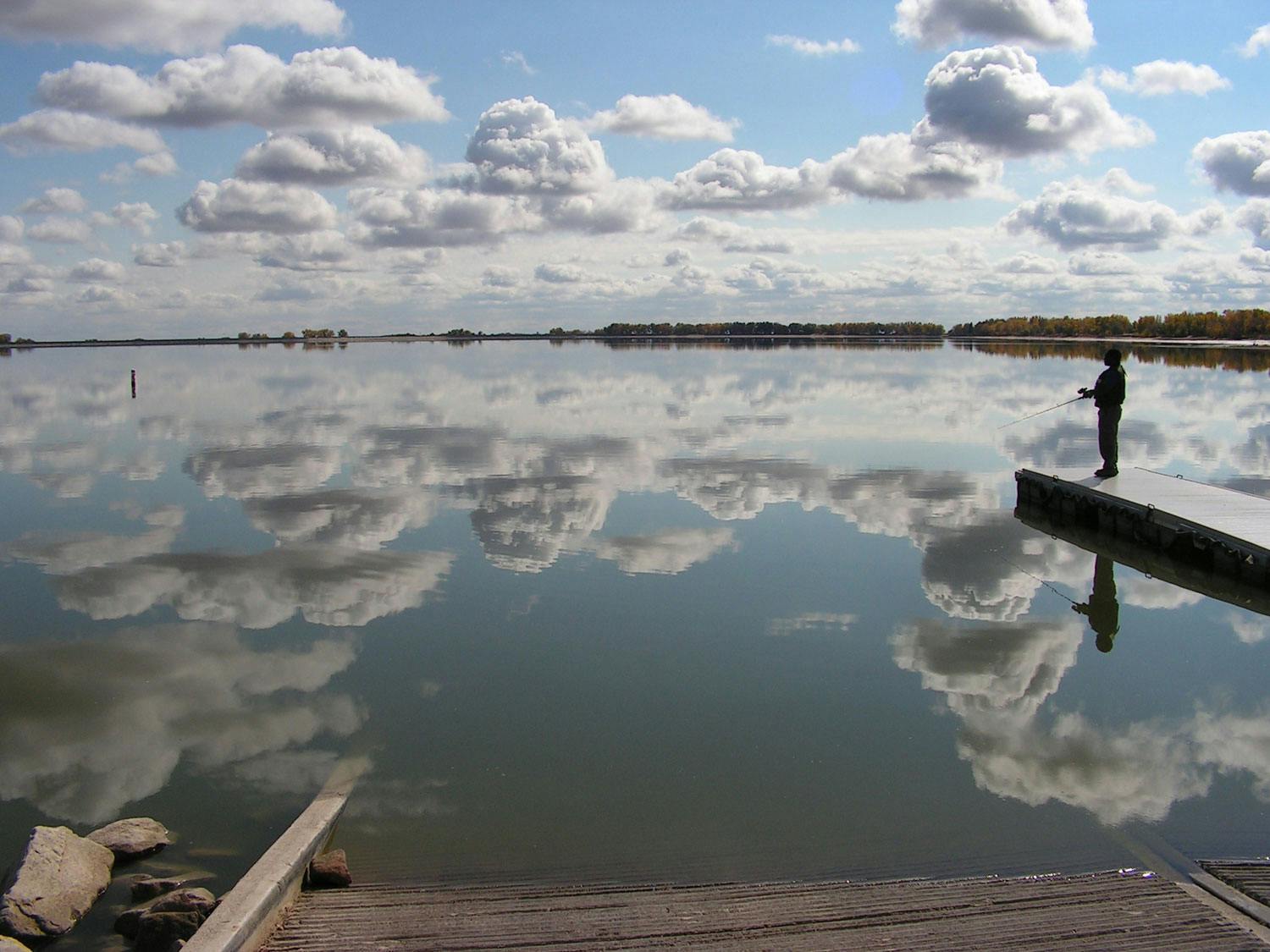 Cotton-ball clouds in the sky are brilliantly reflected in the lake's still waters. A fisher stands on a pier and casts their line