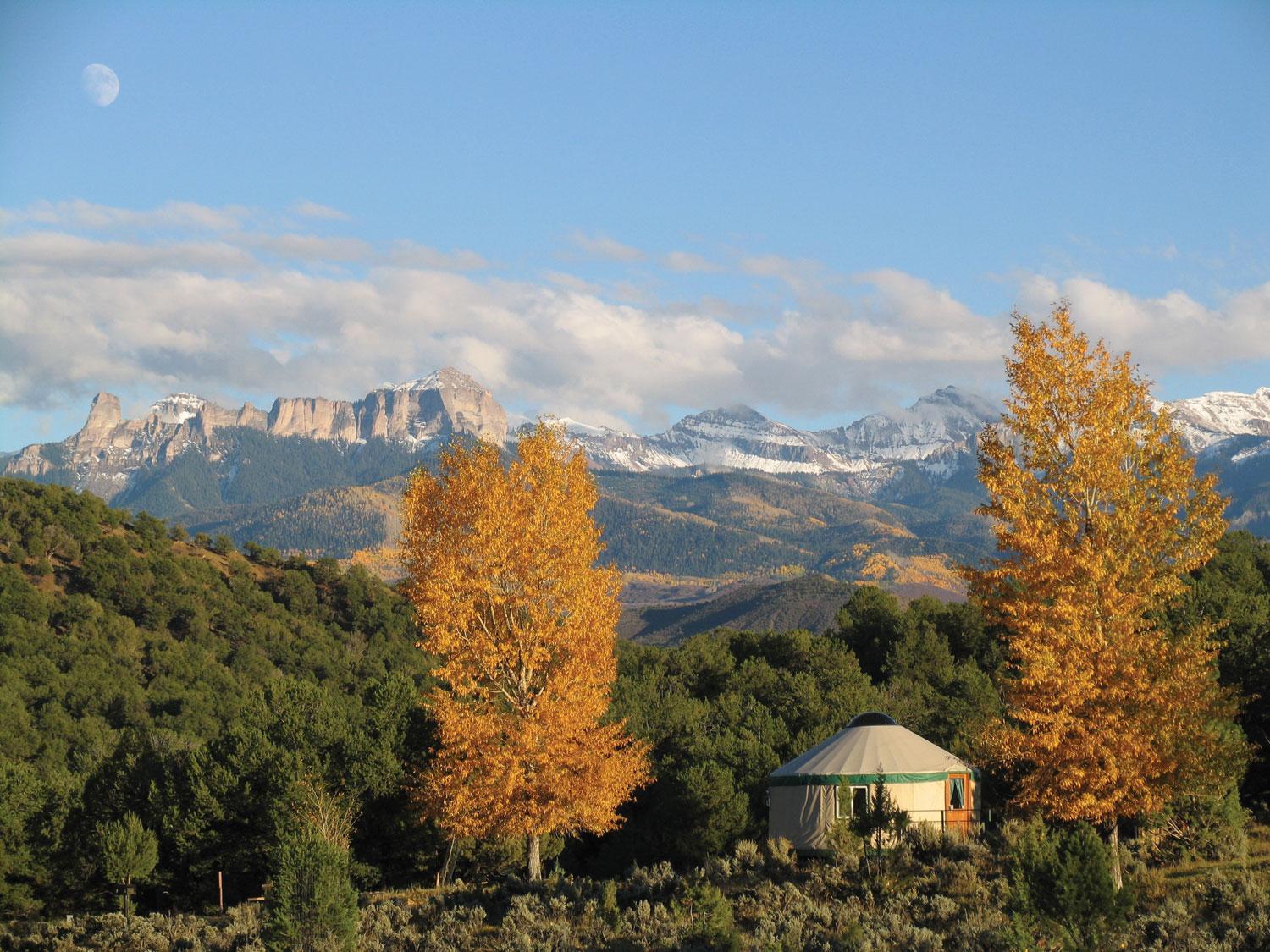 Between two trees with the orange leaves of fall, a circular yurt is nestled beneath a mountain range