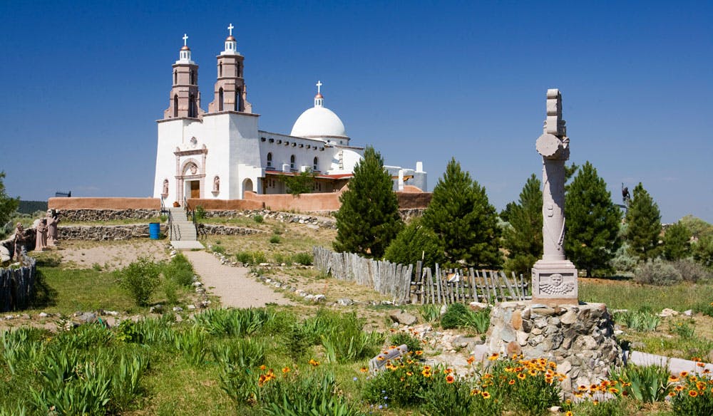 A paved walkway leads up to a white stucco church with twin spires and a dome