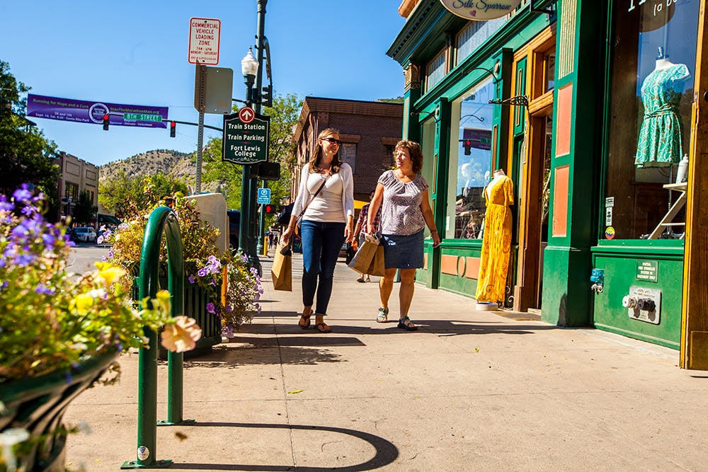 Two people walk down a sidewalk that has flower bushes and a bike rack on the left. On the right a green storefront with a mannequin wearing a yellow dress sit under a blue sky.