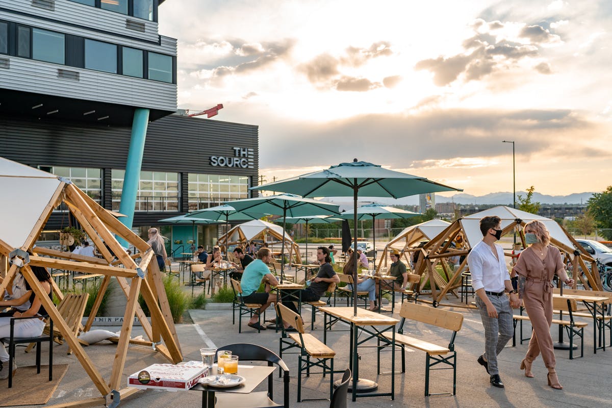 A patio at The Source showcasing a light yellow sky with views of the Front Range. The picnic tables have light-blue umbrellas.