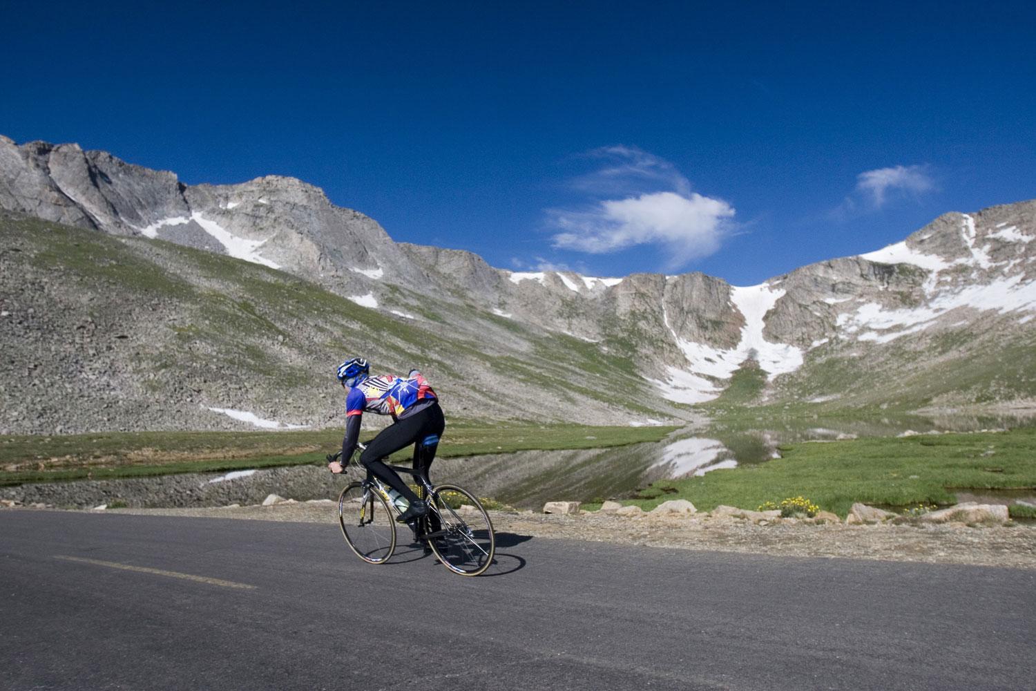 A cyclist zooms up a roadway under bright blue skies and a grassy meadow and a mountian ridge with a trace of snow