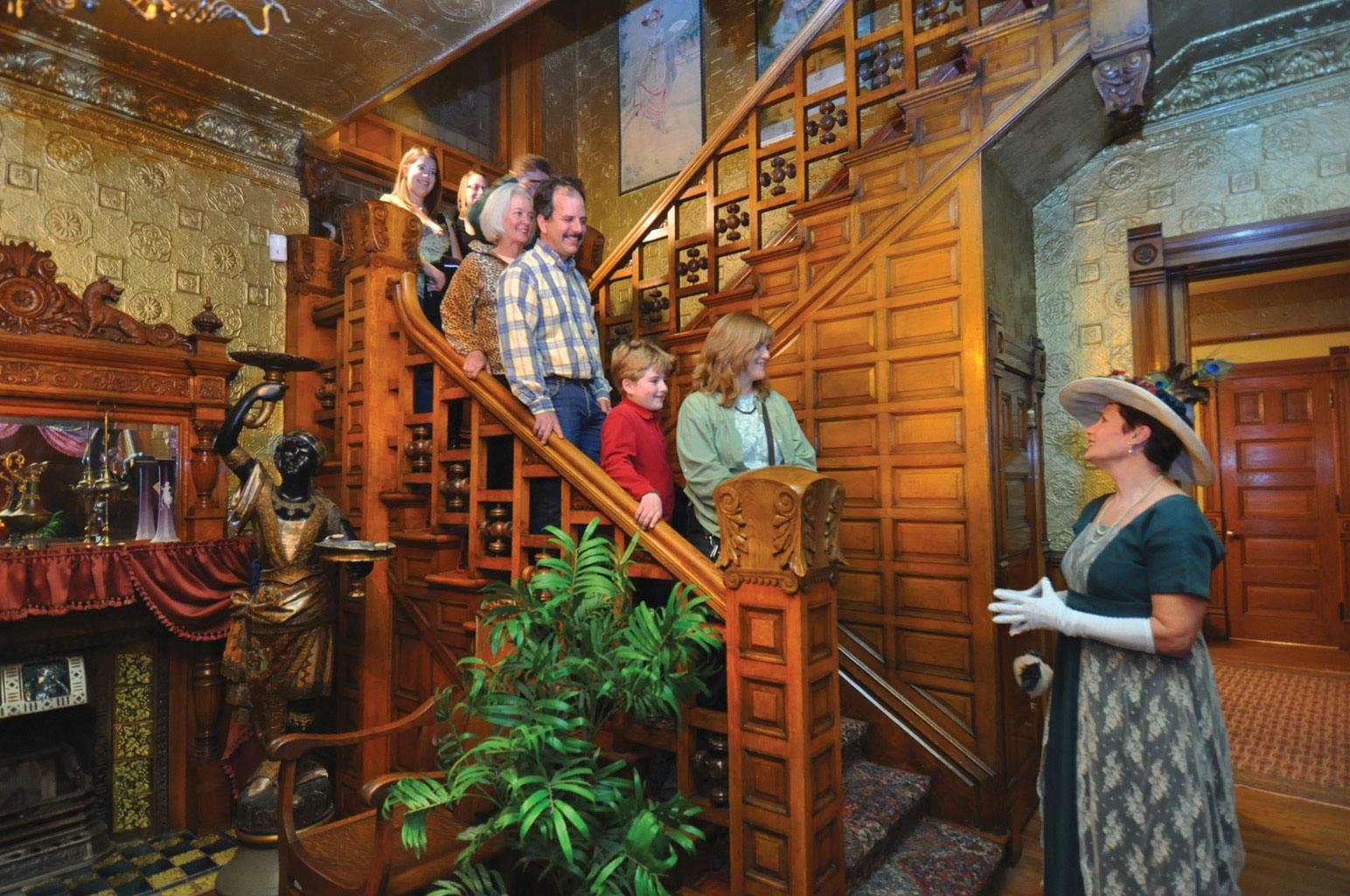 A tour group is stopped on an ornately carved wood staircase, listening to a period actor at the Molly Brown House Museum in Denver.