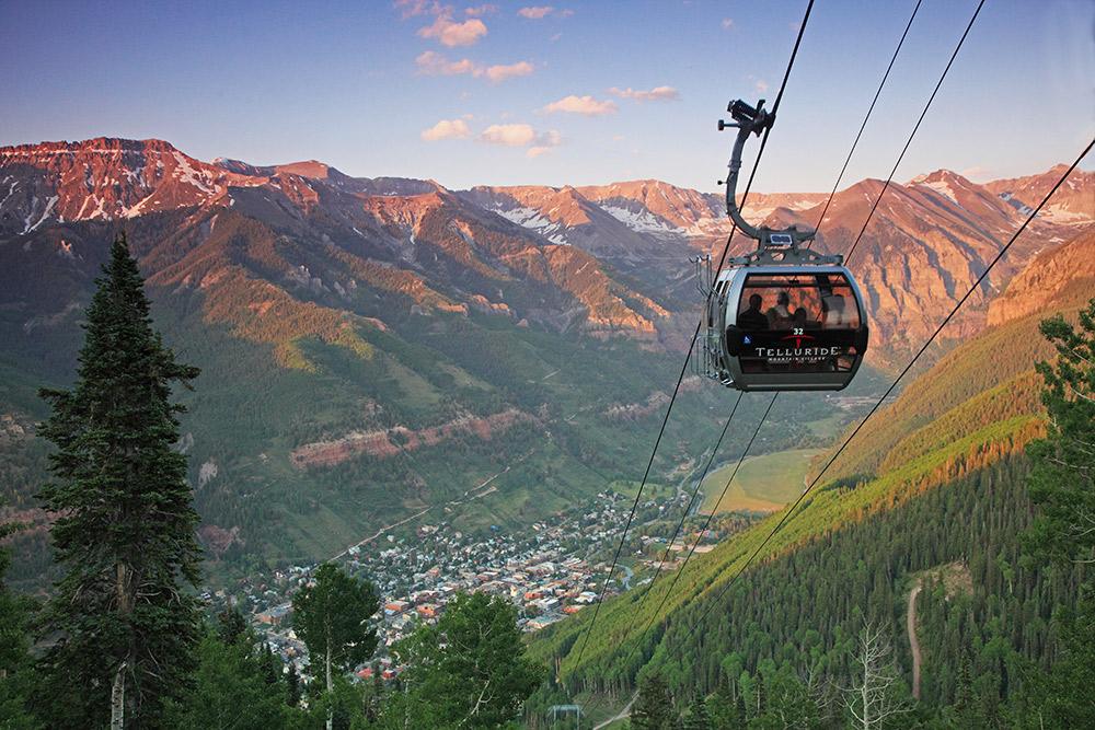 A gondola labeled Telluride makes it's way up the mountain. In the background the town and surrounding mountain peaks are cast in a dusky glow.