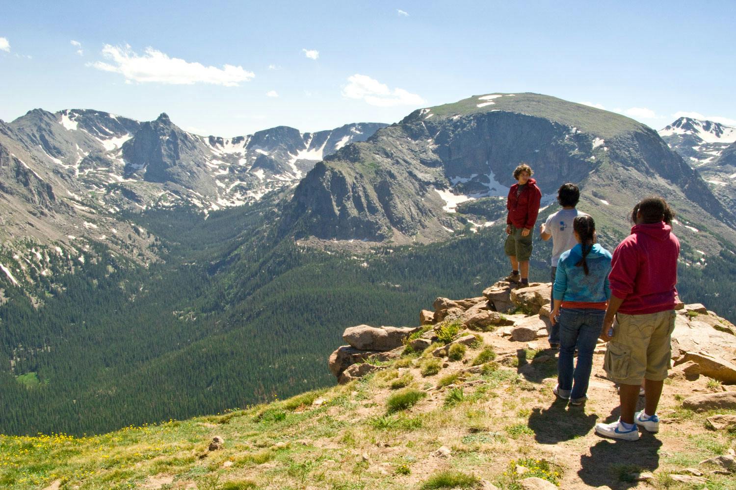 A group of teens stands around a rocky scenic overlook to admire the expansive, lush forested valley below and majestic mountains with pockets of spring snow in Colorado.