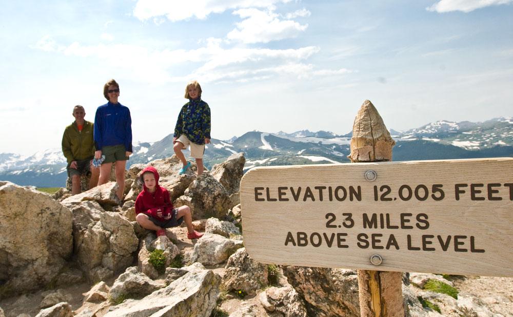 A family of four poses on rocks with mountains in the distance. A sign reads "Elevation 12,005 feet, 2.3 miles above sea level."