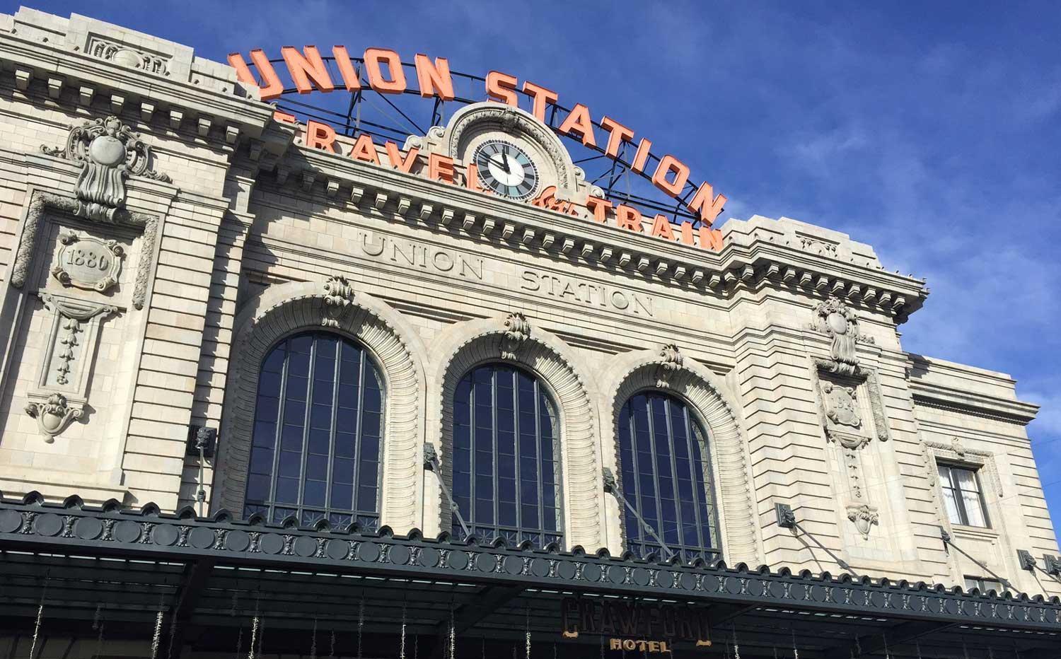 A tall gray-stone building is set against a dark, cloudy sky in Denver, Colorado. Red lettering centered around a clock says, "Union Station Travel by Train."