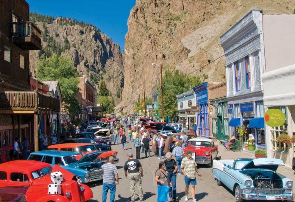 An antique car show takes place in Creede's historic downtown. There are people mingling under a blue sky with rocky mesa mountains in the background.