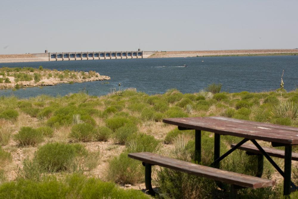 A wooden picnic table sits among green bushes on the shores of a windswept lake; a dam is in the distance